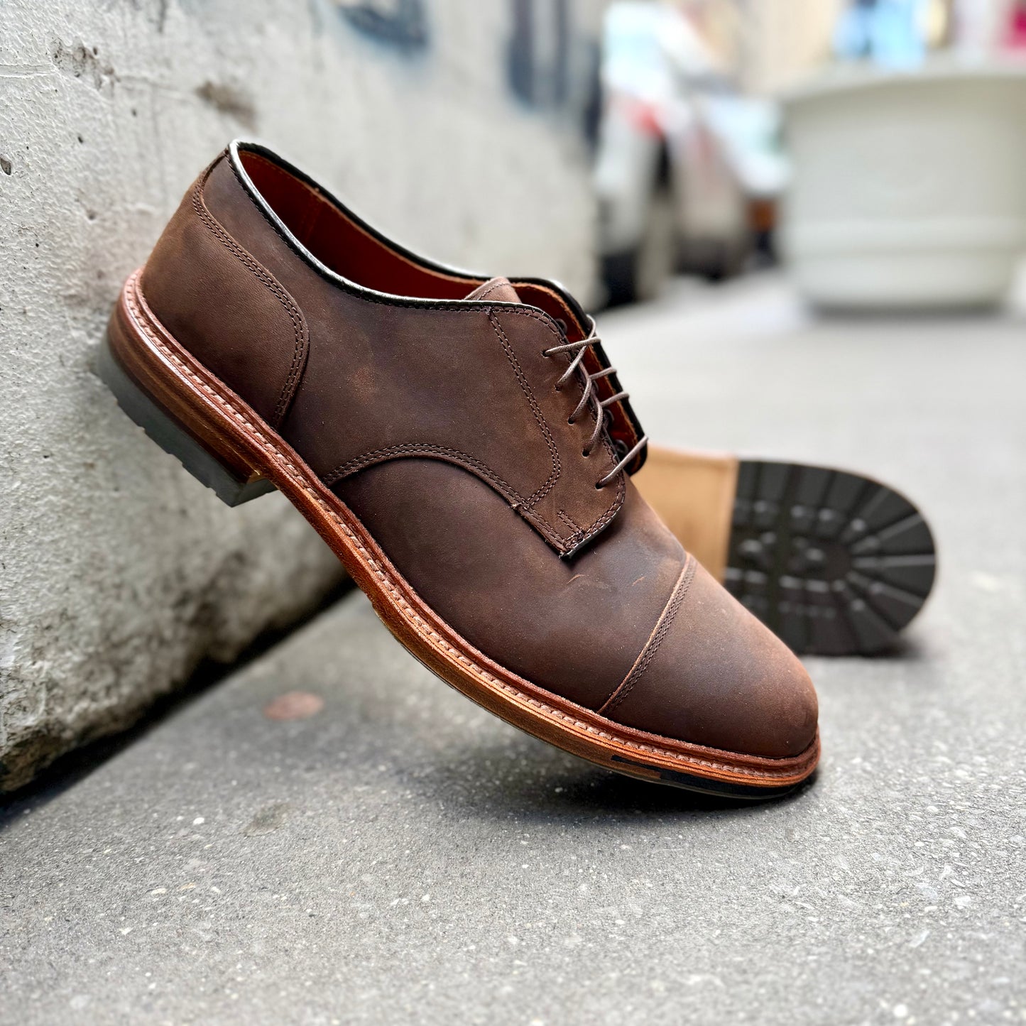 A pair of Alden Madison D4519C - Tobacco Chamois Cap Toe Bluchers with laces and polished wooden soles sits on a concrete sidewalk against a textured wall, with a blurred city street in the background.