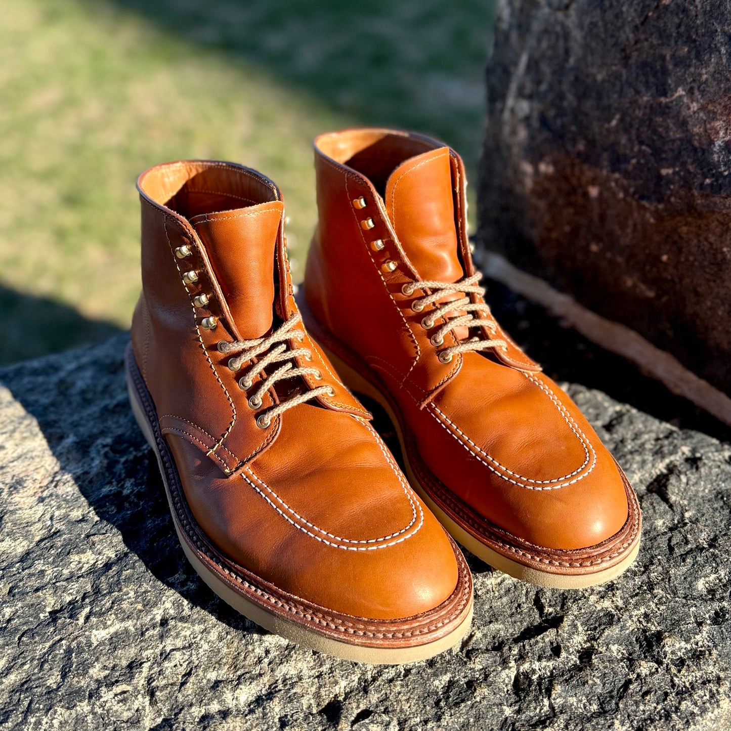 A pair of Alden Madisons Sherwood Indy Boots (40311H) in Saddle Calf leather, featuring tan soles, rests on a large rock. Sunlight illuminates their texture and shine against the grassy backdrop.
