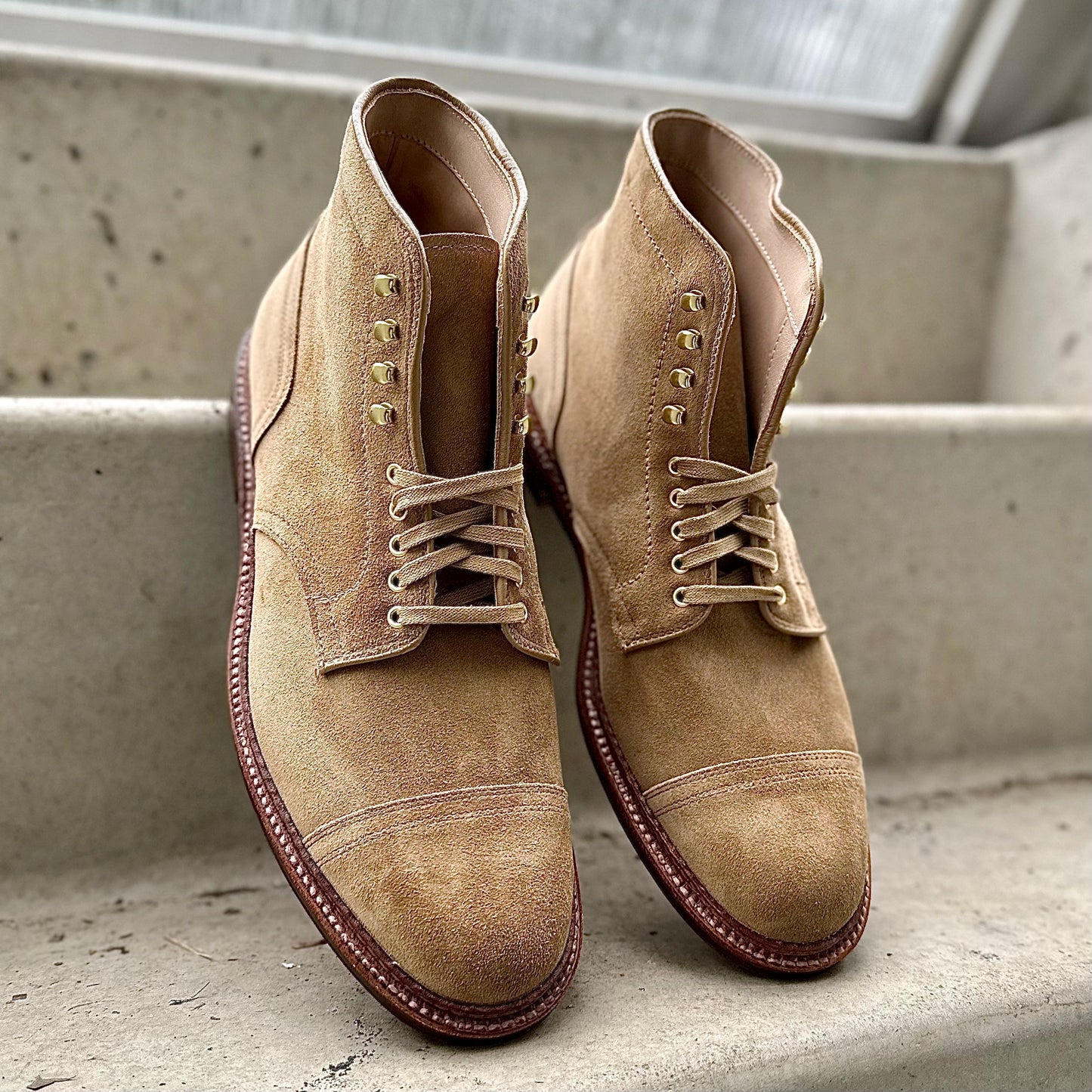 A pair of Alden Madisons D2685H - 2/2 Cap Toe Boots in Tan Suede, featuring brown soles and metal eyelets, sits on concrete steps. The classic boots showcase visible stitching against an out-of-focus background.