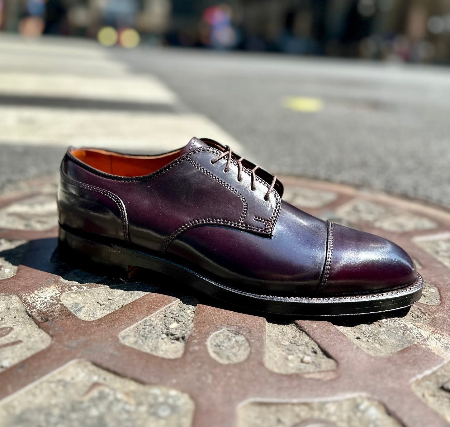 The Alden Madison 2160 - Cap Toe Blucher in Color 8 Shell Cordovan rests on a decorative manhole cover in a sunlit urban street, with a blurred city background.
