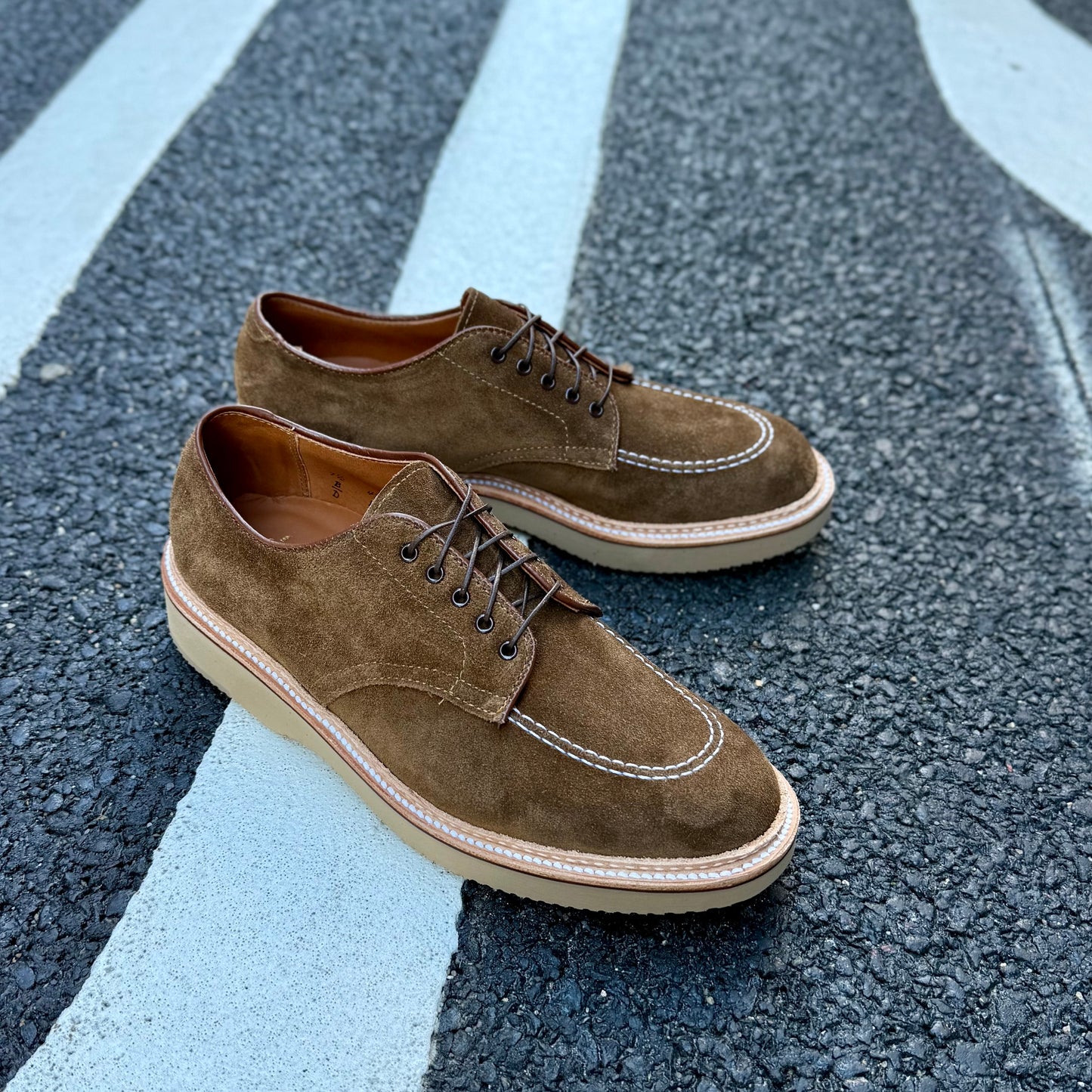 The ALDEN SHOES M7601Y, a snuff suede Indy shoe with wedge, features brown suede with light soles, white stitching, and dark laces. Placed on a textured gray surface with white lines, these shoes are crafted on the Trubalance last for style and comfort.