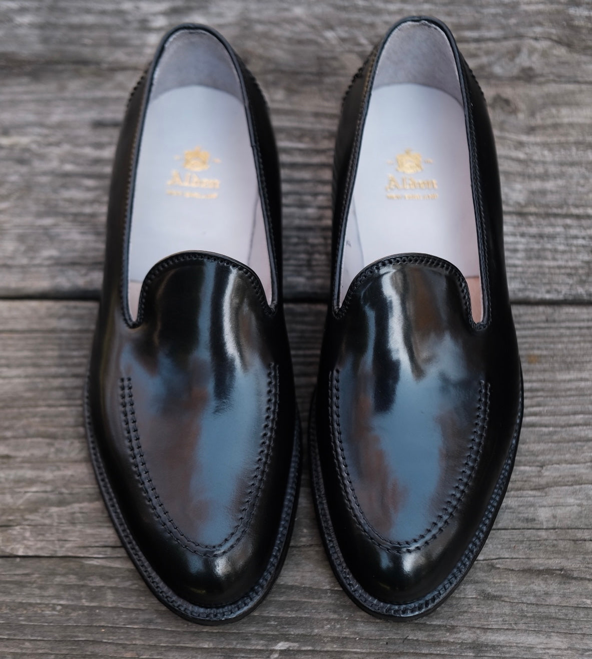The Alden Madison D2117 - Vanderbilt Slip On in Black Shell Cordovan loafers rest side by side on a weathered wooden surface, featuring polished black leather and visible stitching. The insoles are light-colored with gold text imprinted on them.