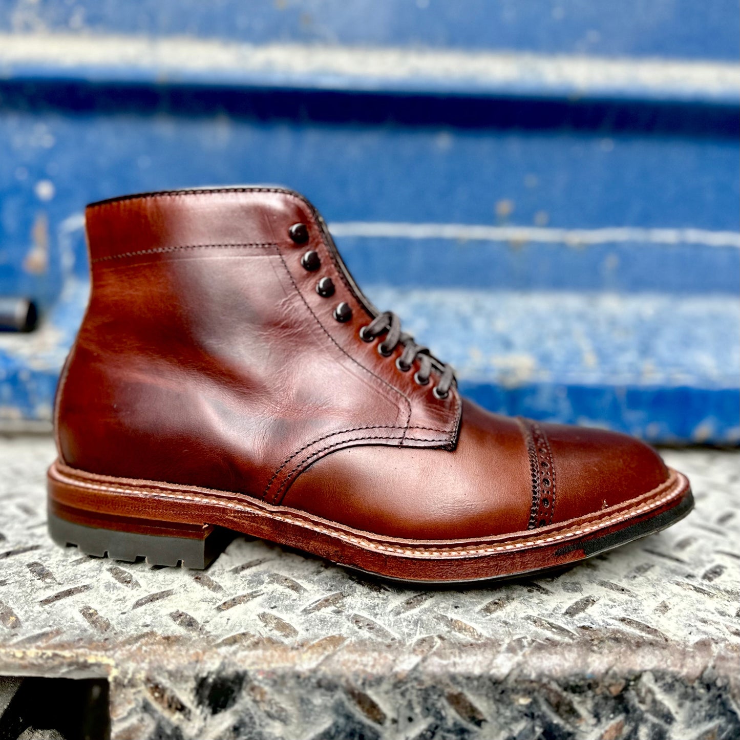 The Alden Madison 44924HC Jumper Boot in Brown Chromexcel, featuring decorative stitching and speed hooks, is displayed on a textured metal surface against a blue industrial background.