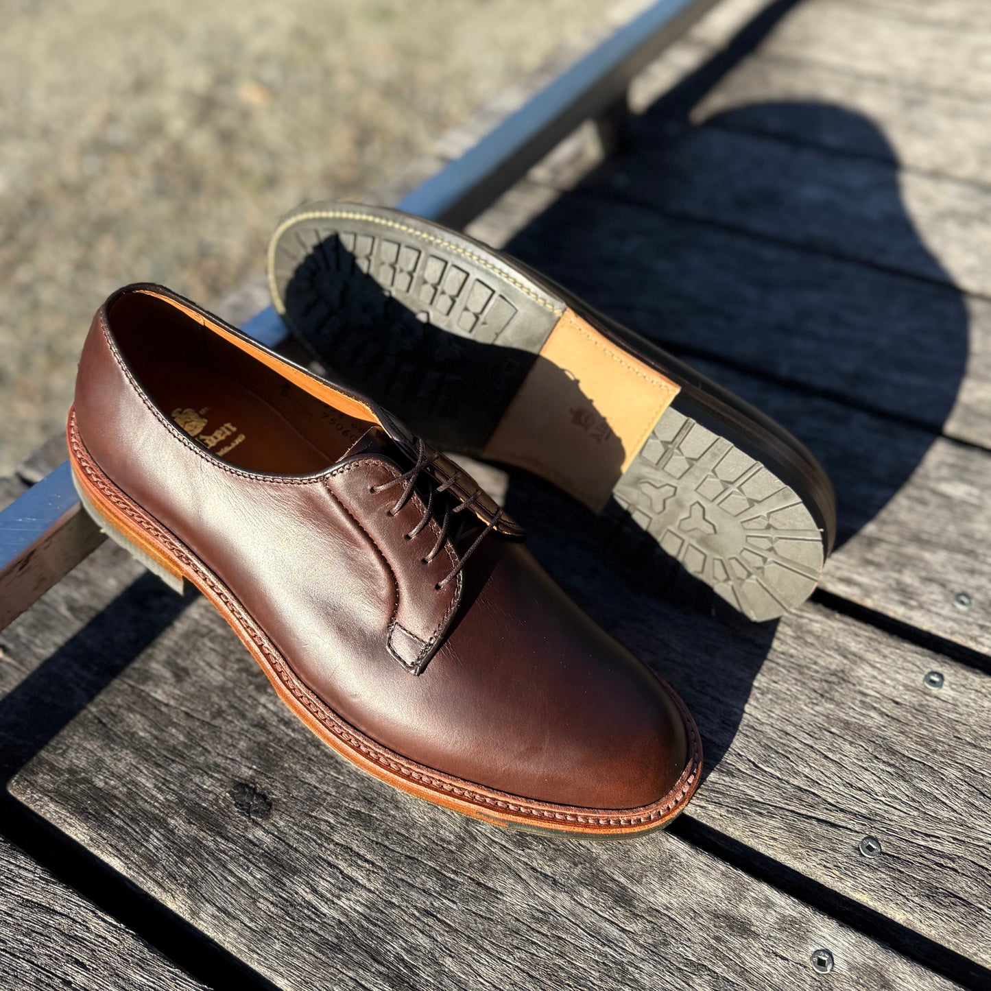 A pair of ALDEN SHOES 9506C Plain Toe Blucher in Brown Chromexcel with Commando rubber soles is shown on wood in sunlight; one shoe stands upright, the other (on the Barrie Last) is flipped to display its tread.