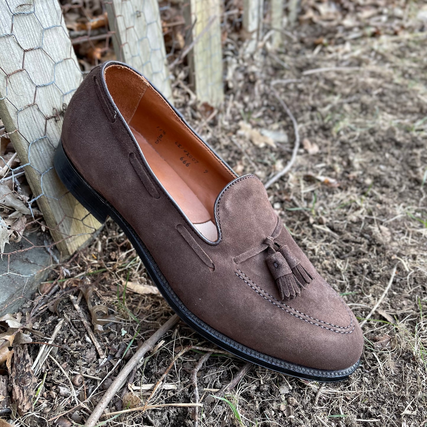 The Alden Madison 666 Tassel Loafer in Brown Suede sits by a wooden fence on grass and dry leaves. Crafted on the Aberdeen Last, it boasts a black leather sole and intricate edge stitching, exuding timeless elegance.