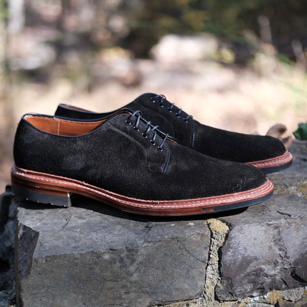 A pair of Alden Madisons D1426C - Plain Toe Blucher in Earth Reverse Chamois with Commando Soles and black laces sits on a stone surface, set against a blurred natural backdrop.