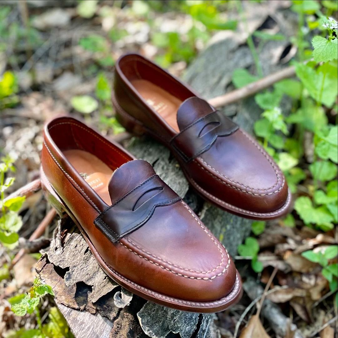 The Alden Madison 17831F - Unlined Leisure Hand Sewn loafer in Brown Chromexcel leather, featuring stitching detail and a classic leather sole, rests on weathered wood amidst green foliage and fallen leaves.