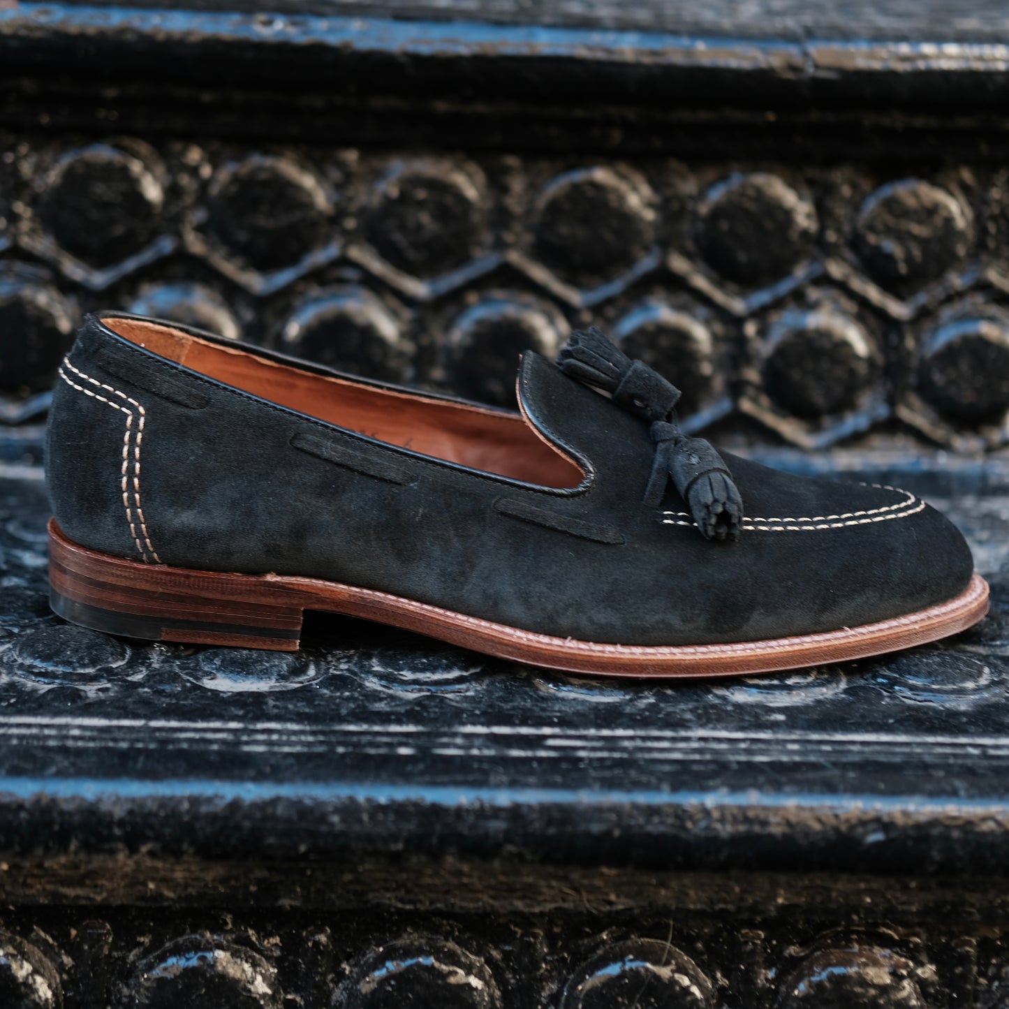 The Alden Madison D0106 Black Suede Tassel Loafer, crafted on the Aberdeen Last with white stitching and a brown leather sole, is shown on a textured black metal step.