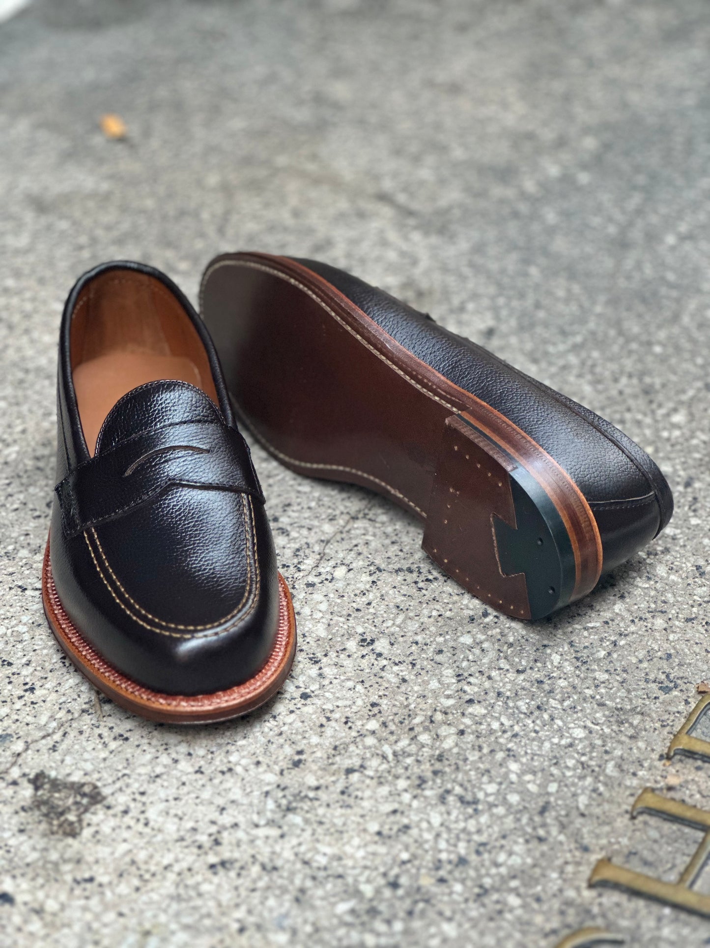 A pair of Alden Madisons 62456F Penny Loafers in Brown Regina Grain, featuring tan stitching, is displayed on a textured concrete surface. One shoe is upright to show its top, while the other reveals the Flex Leather Sole.