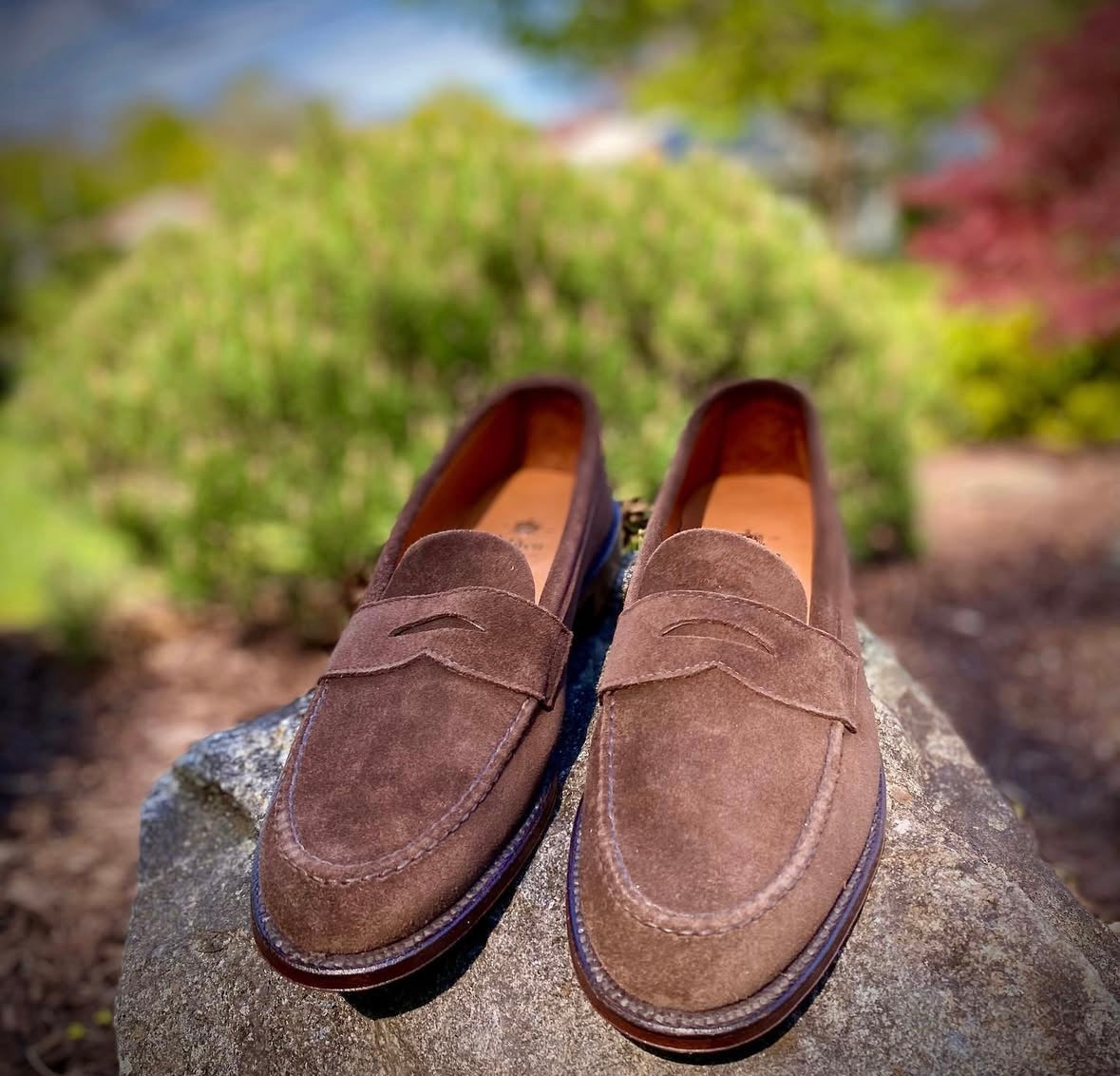 The Alden Madison 6245F - Unlined Leisure Hand Sewn in Brown Suede with Flex Leather Sole is displayed outdoors on a large rock, set against a blurred backdrop of greenery and plants.