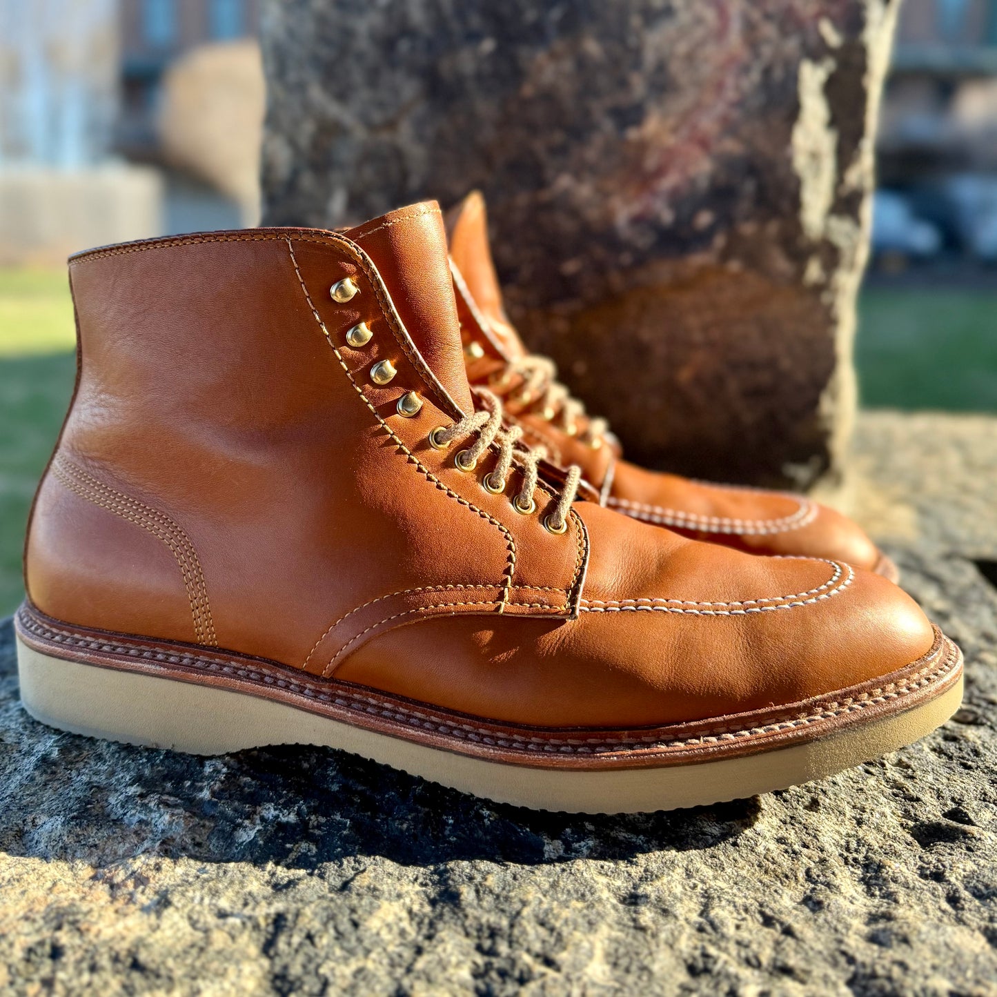 A pair of Alden Madisons 40311H Sherwood Indy Boots in Saddle Calf leather, featuring white stitching and brass eyelets, rests outdoors on a rock. Lit warmly by sunlight, their impeccable design on the Trubalance last is highlighted by a light-colored sole.