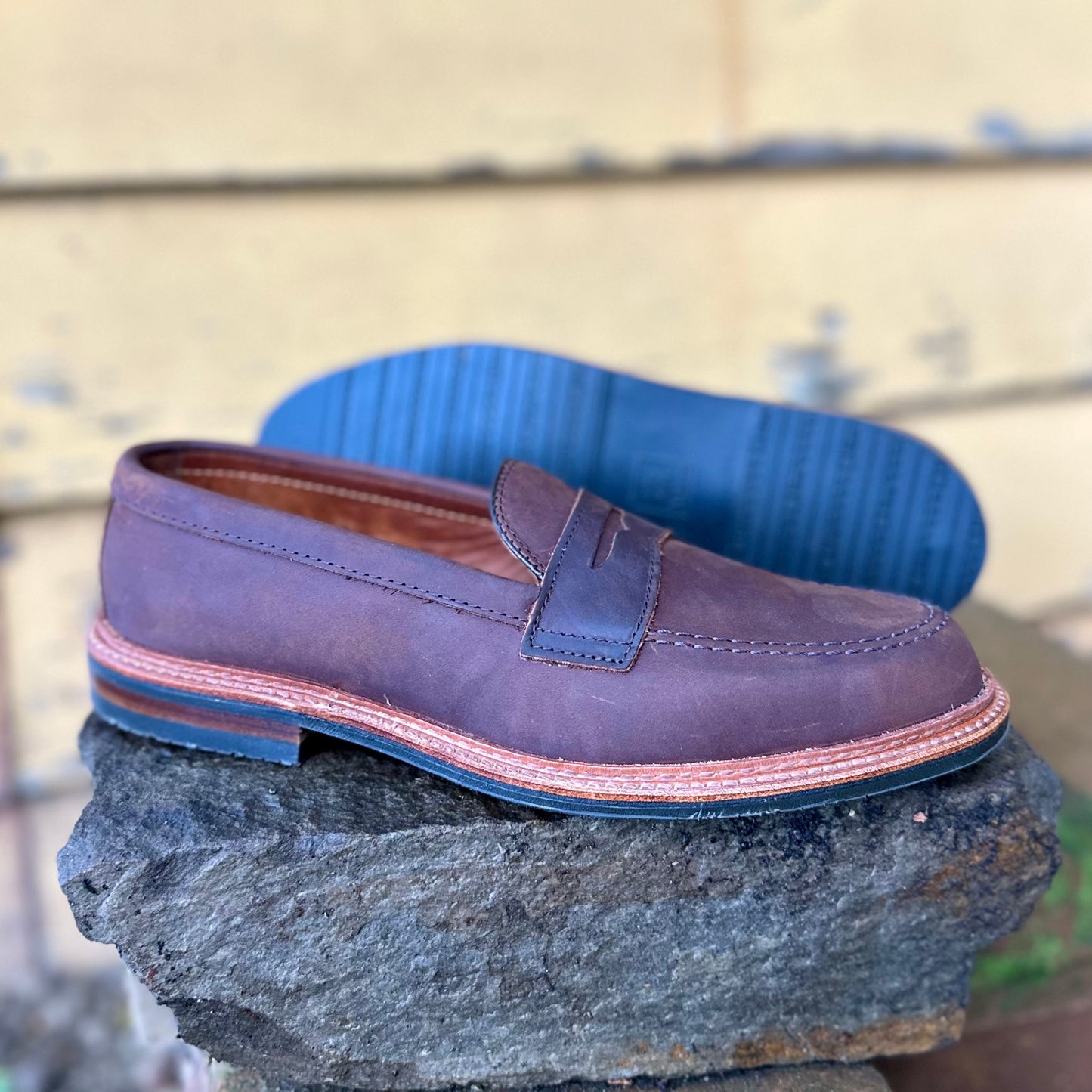 The D2216L - Smooth Tobacco Chamois Penny Loafer by Alden Madison, featuring brown leather and a blue rubber sole with stitching details, is showcased on a rough stone surface. A faded wooden wall in the background enhances the shoes classic design with an antique edge.