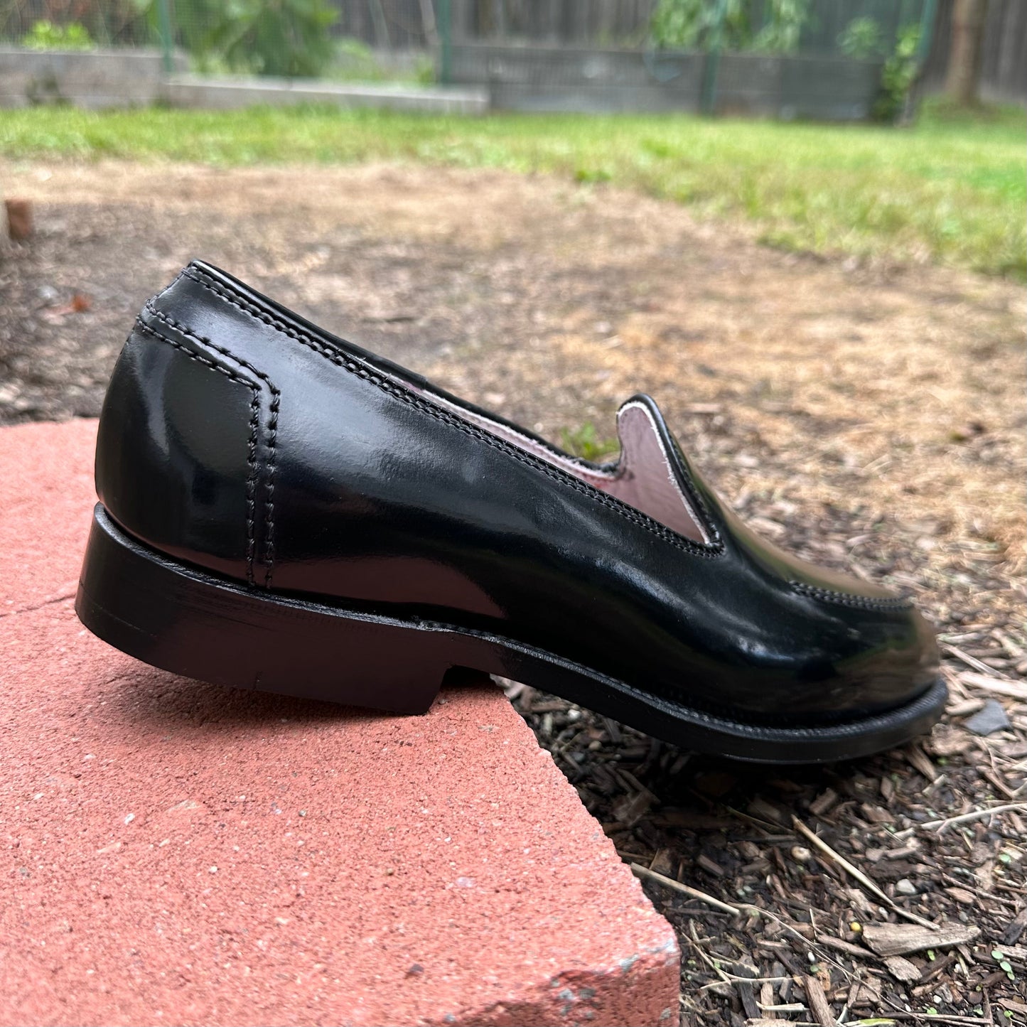 The D2117 - Vanderbilt Slip On by Alden Madison, crafted in black shell cordovan, rests on a red brick with a grassy garden behind. From the side, its sleek design, shiny surface, and elegant Brooks heel foxing stand out.