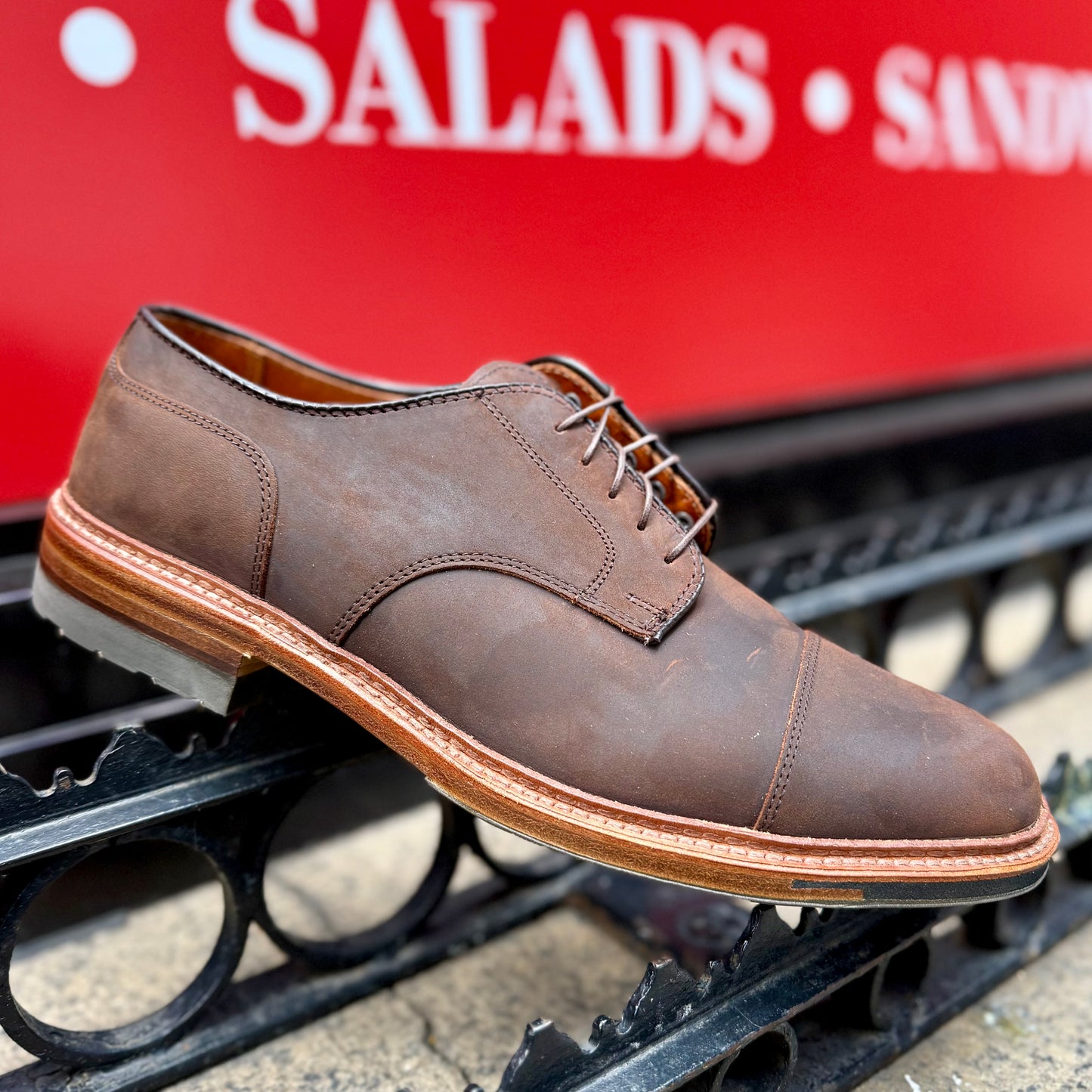 Aldens Madisons D4519C - Tobacco Chamois Cap Toe Blucher, a brown leather shoe with laces and a light brown commando sole, rests on a wrought iron fence against a backdrop of a red sign showing SALADS and SANDW.