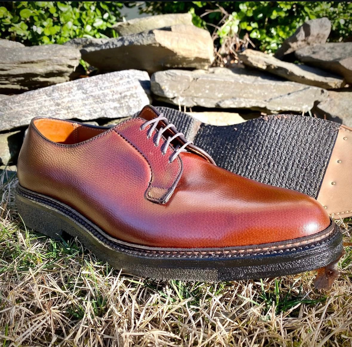 The Alden Madison 947 - All Weather Walker in Brown Alpine Grain sits on dry grass with a black crepe sole and white laces. A stone wall and greenery are in the background, and another shoe is partially visible behind it.