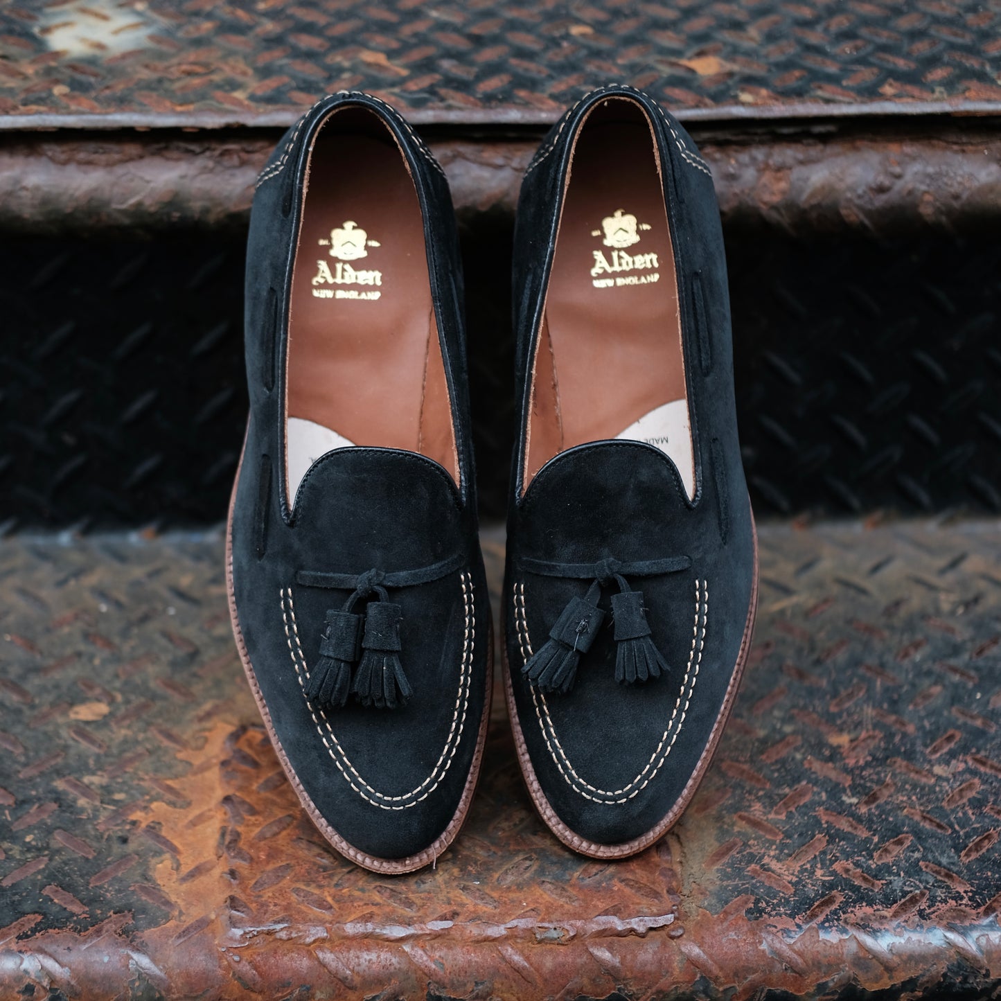 Alden Madison D0106 Black Suede Tassel Loafers, with white stitching and brown leather interiors featuring gold branding, are displayed side by side on a rusty, textured metal staircase.