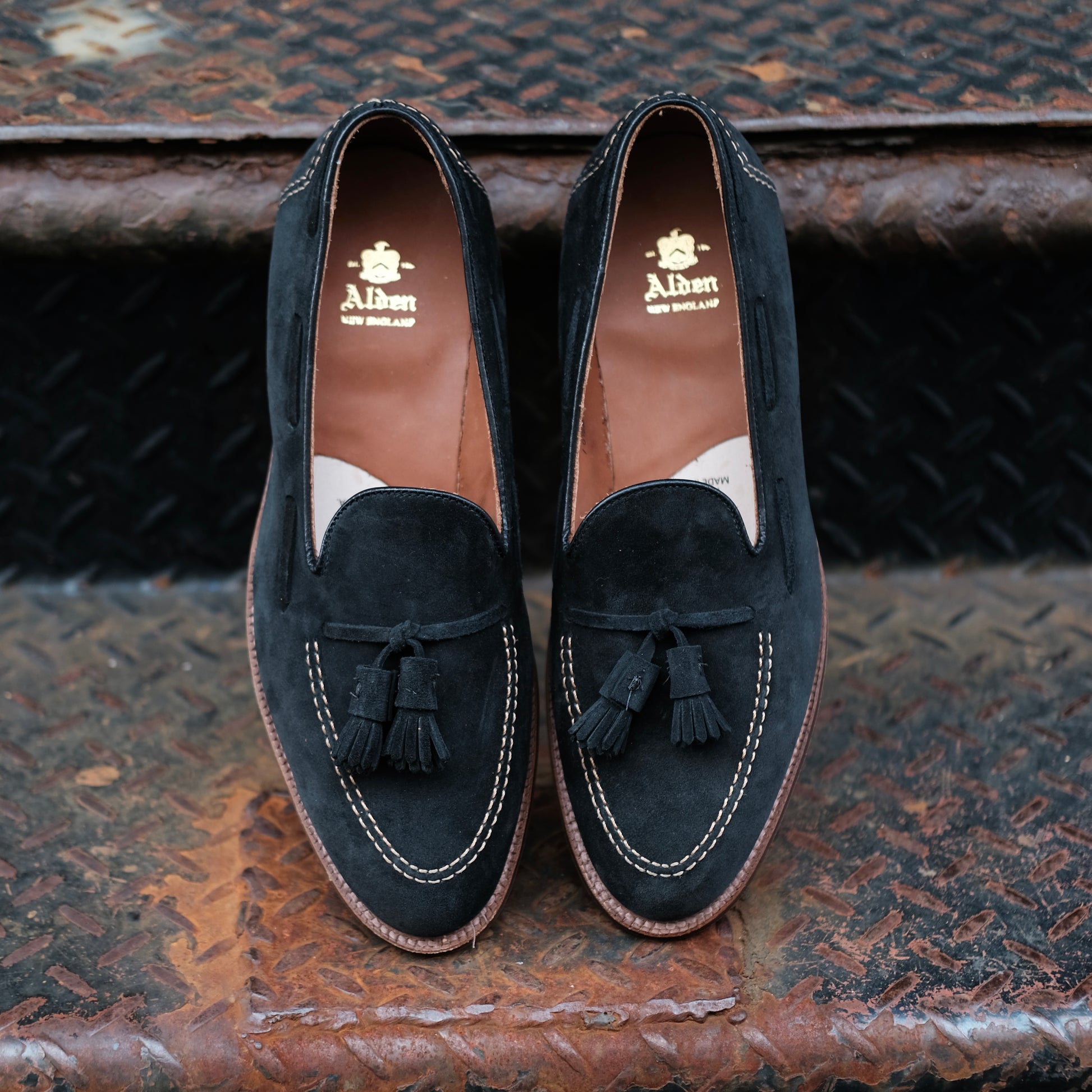 Alden Madison D0106 Black Suede Tassel Loafers, with white stitching and brown leather interiors featuring gold branding, are displayed side by side on a rusty, textured metal staircase.