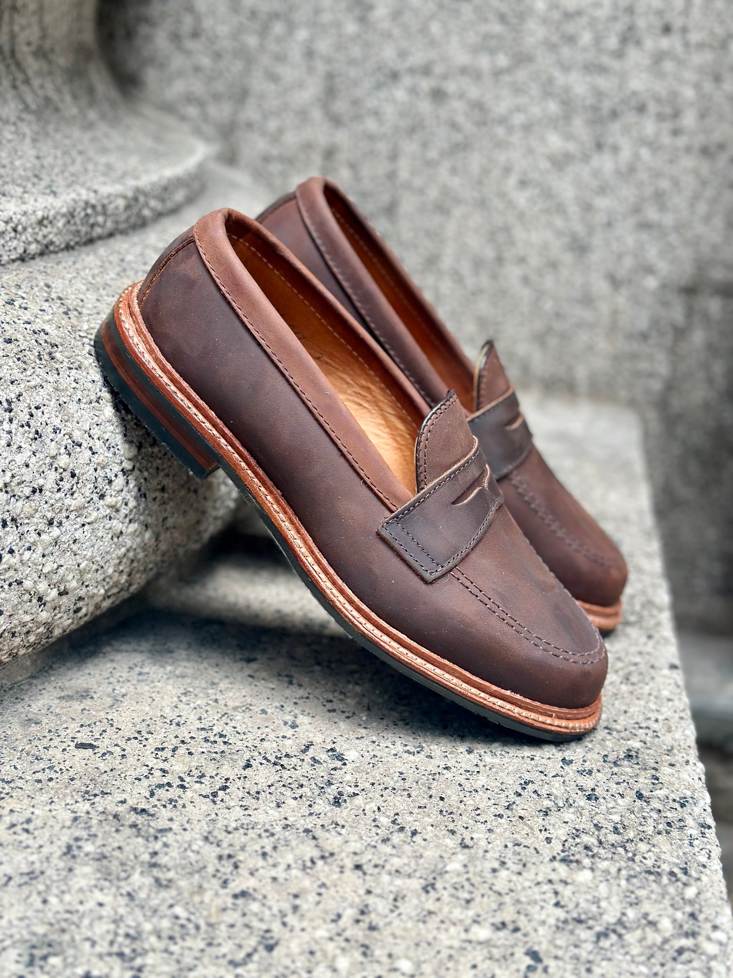 The Alden Madison D2216L - Smooth Tobacco Chamois Penny Loafer, featuring visible stitching and a durable rubber sole, rests on a gray stone surface.
