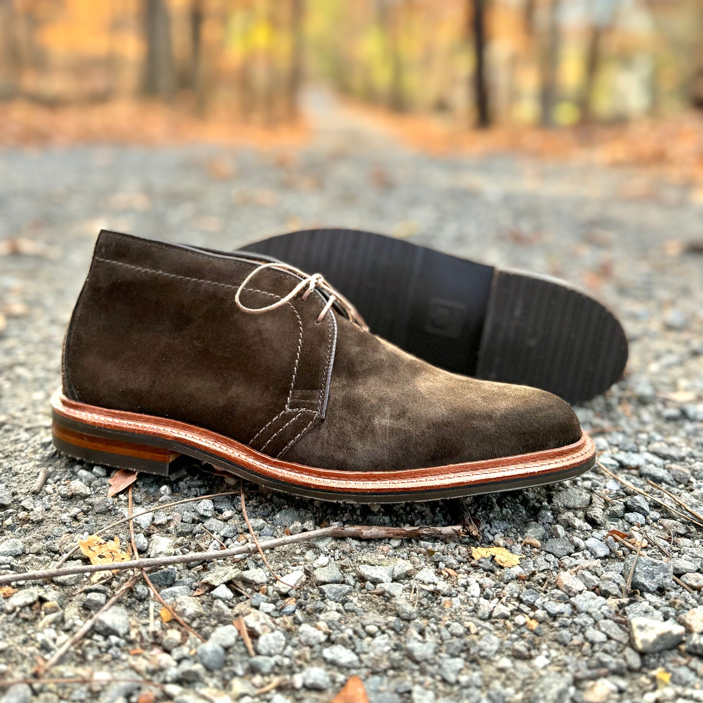 The D1712L Chukka Boot from Alden Madison, crafted in loden suede on the Barrie Last, features light brown laces. One boot is upright and the other lies sideways on a gravel path amid autumn leaves, highlighting its reverse welt and textured L3 sole.