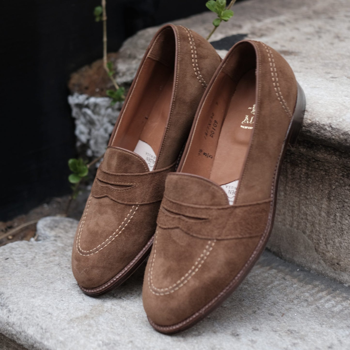 The Alden Madison D0109 - Full Strap Loafer in Snuff Suede (Deposit) features white stitching and a leather outsole, resting on outdoor stone steps amid small green plants.