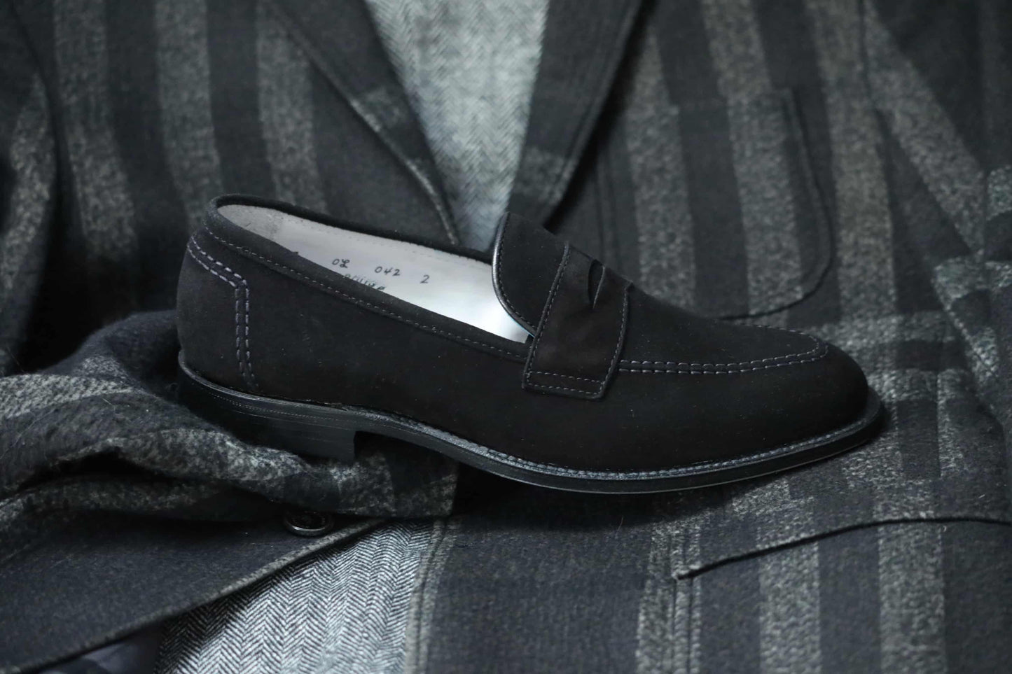 The Alden Madison D2229 “Stuyvesant” Madison Penny in black suede, with white stitching and a leather sole, is displayed atop a dark striped suit jacket, revealing its herringbone-patterned sleeve beneath.