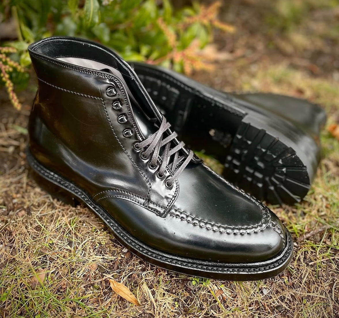 The Alden Madison 40219HC - NST Tanker Boot in Black Shell Cordovan, with visible stitching and a sturdy Commando Sole, is shown on grass—one boot upright, the other lying on its side amid greenery.