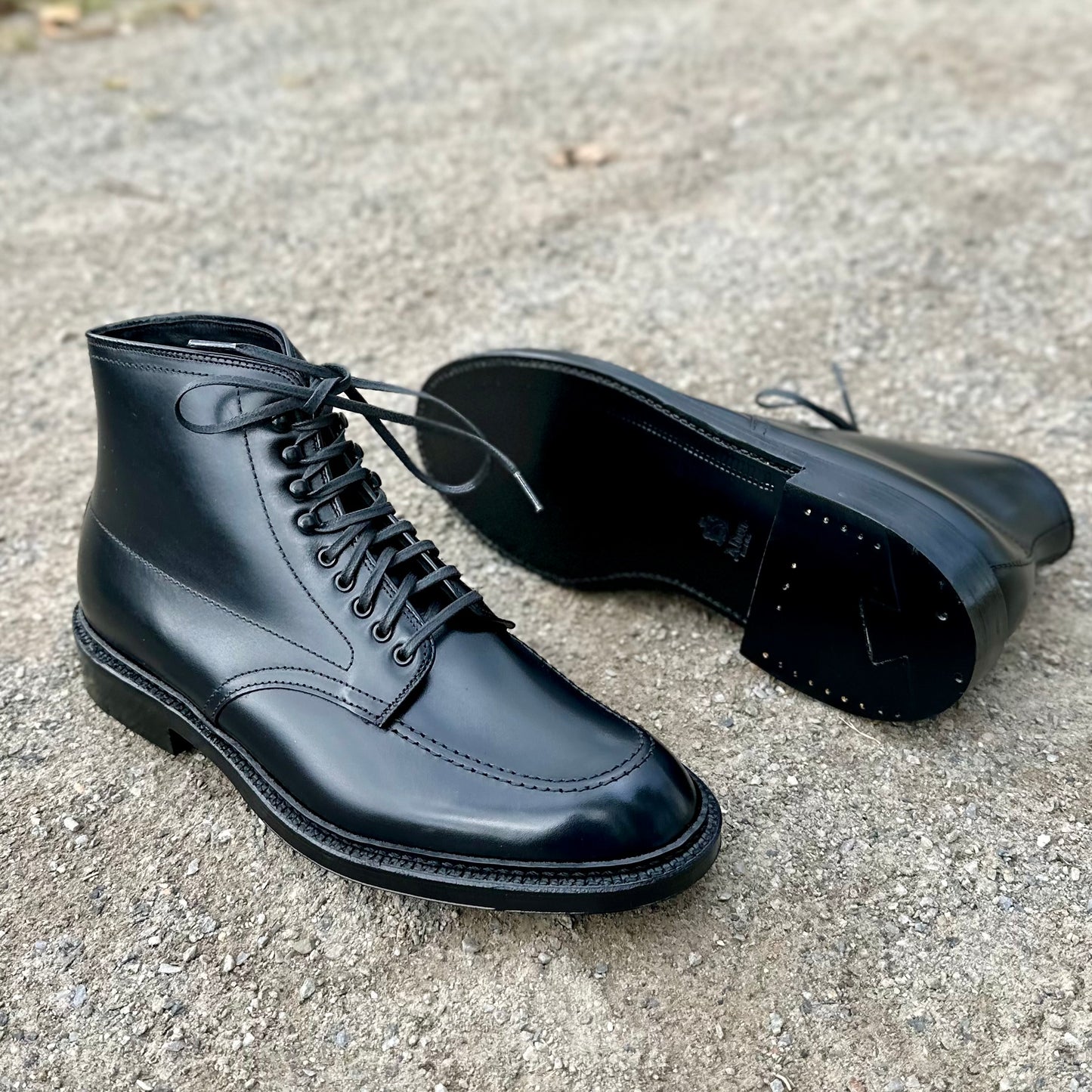 A pair of Alden Madisons D2941H - Black Calf Indy Boots with laces, featuring a double leather sole, is showcased on a textured gray surface. The polished boots highlight intricate stitching; one stands upright while the other lies on its side.