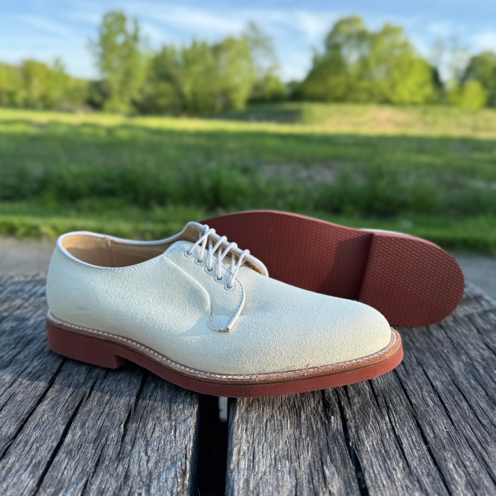 The Alden Madison 9448 - White Buck in Ivory Suede, featuring brown soles, is showcased outdoors on a rustic wooden surface with green grass and trees softly blurred in the background.
