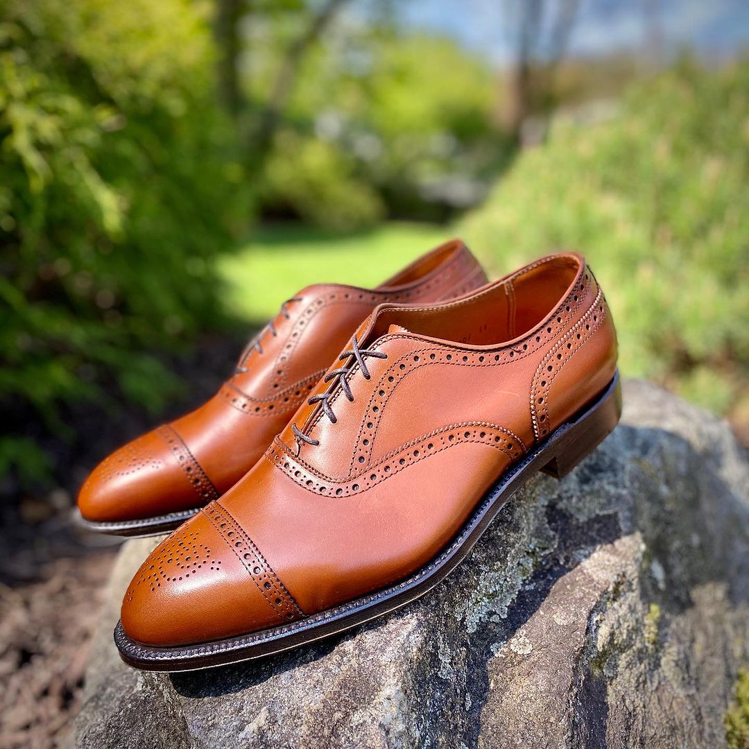 The Alden Madison 911 Burnished Tan Medallion Cap Toe Bal shoes, featuring decorative brogue detailing and crafted on the Hampton Last with a fine leather sole, rest on a rock surrounded by lush green foliage under a sunny sky.