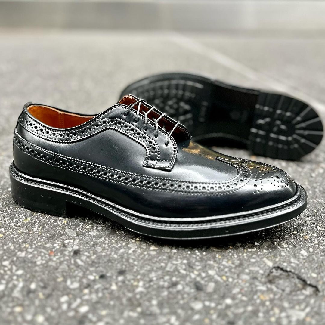 The Alden Madison 9751C Long Wing Blucher in black shell cordovan is showcased on a textured gray surface, featuring intricate brogue detailing. One shoe with a Commando sole stands upright while the Barrie Last model lies on its side, displaying its sole.