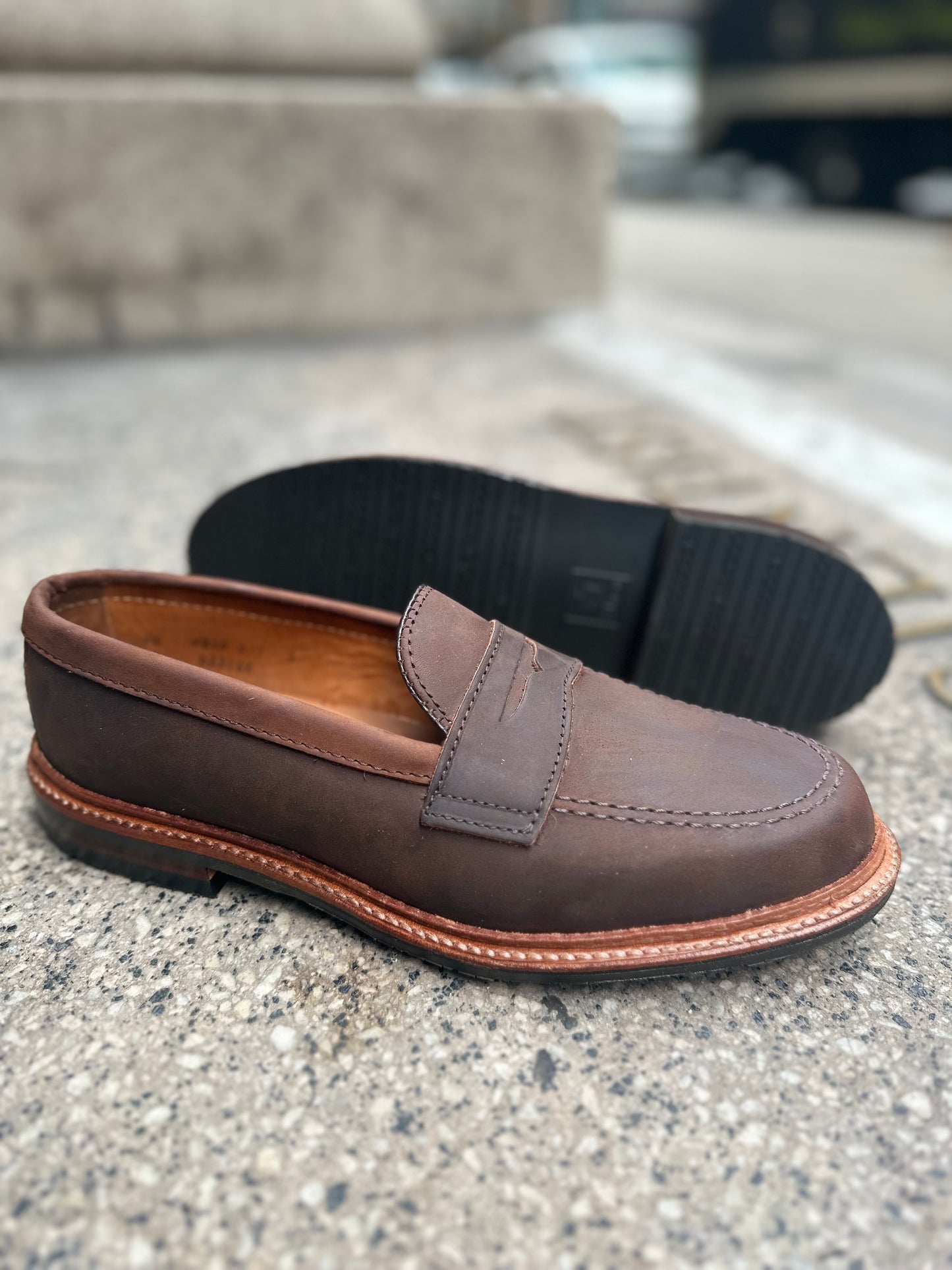 The Alden Madison D2216L - Smooth Tobacco Chamois Penny Loafer, with antique edge and visible stitching on a rubber sole, stands upright on concrete while another lies sideways, revealing its intricate tread pattern.