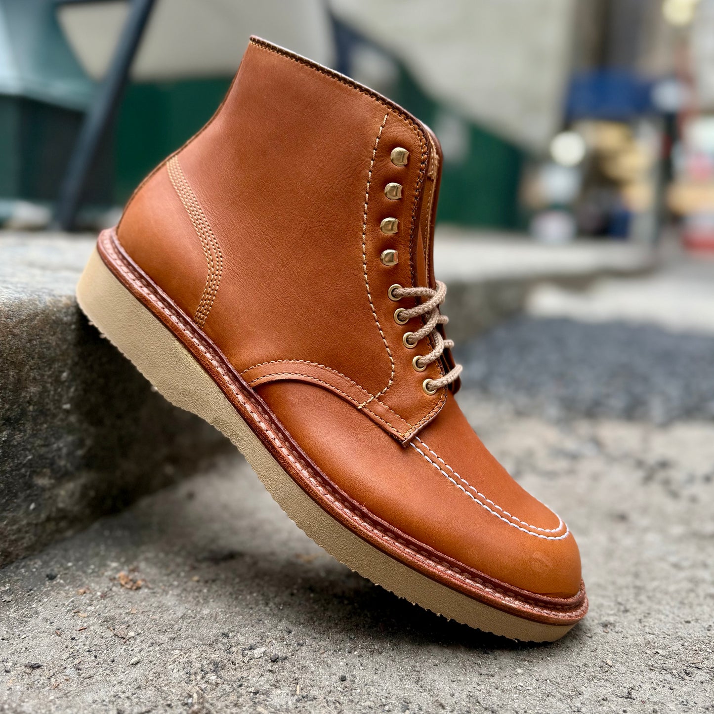 The Alden Madison 40311H Sherwood Indy Boot in saddle calf showcases rugged elegance with a cream wedge sole, tan leather, detailed stitching, and light brown laces against a concrete curb in an urban setting.