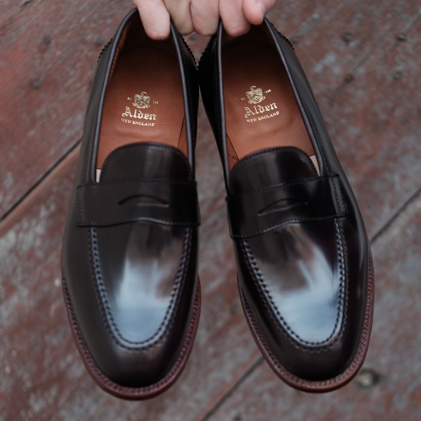 A pair of Alden Madisons 96288 - Madison Penny loafers in polished Color 8 Shell Cordovan, showcasing a visible brand logo on the insoles and elegant antique edging, held over a textured wooden surface.