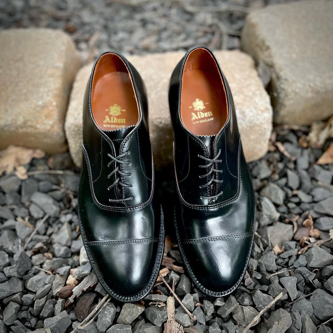 A pair of Alden Madison 9071 - Straight Tip Bal dress shoes in Black Shell Cordovan sit side by side on dark gravel, with two rectangular stone blocks behind them.