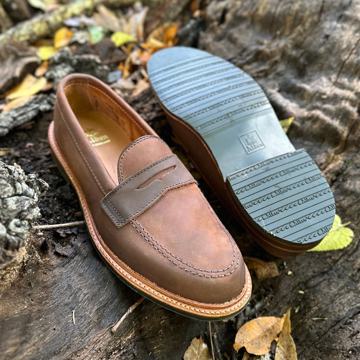 A pair of Alden Madisons D2216L - Smooth Tobacco Chamois Penny Loafers is placed on a wooden surface amid autumn leaves. One shoe stands upright, highlighting its stitched upper and antique edge, while the other is tilted to reveal its patterned rubber sole.