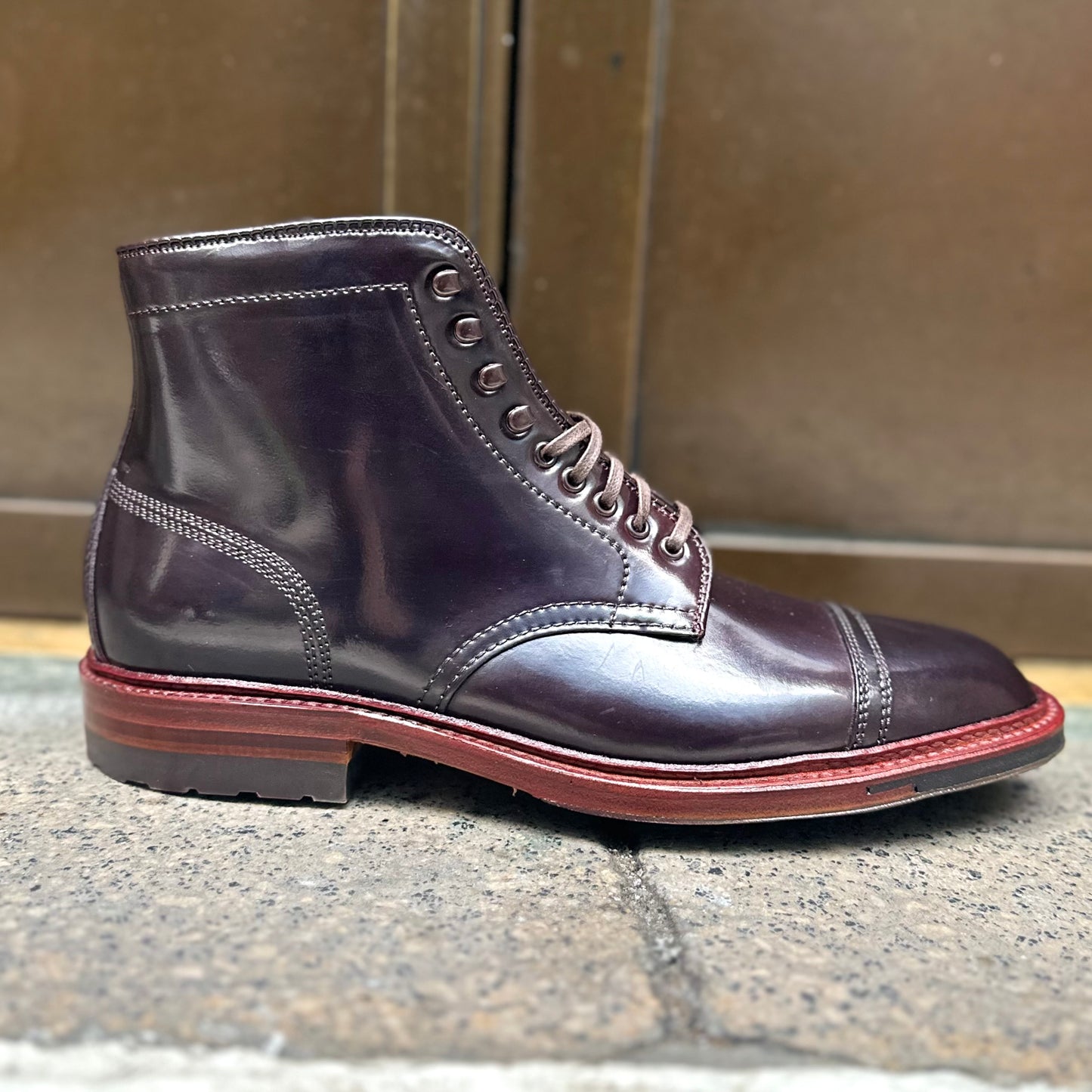 The Alden Madison D2858HC - 2/2 Cap Boot in Color 8 Shell, featuring a polished dark brown leather with laces and a durable red-brown commando sole, is elegantly displayed sideways on a stone surface against a blurred background.