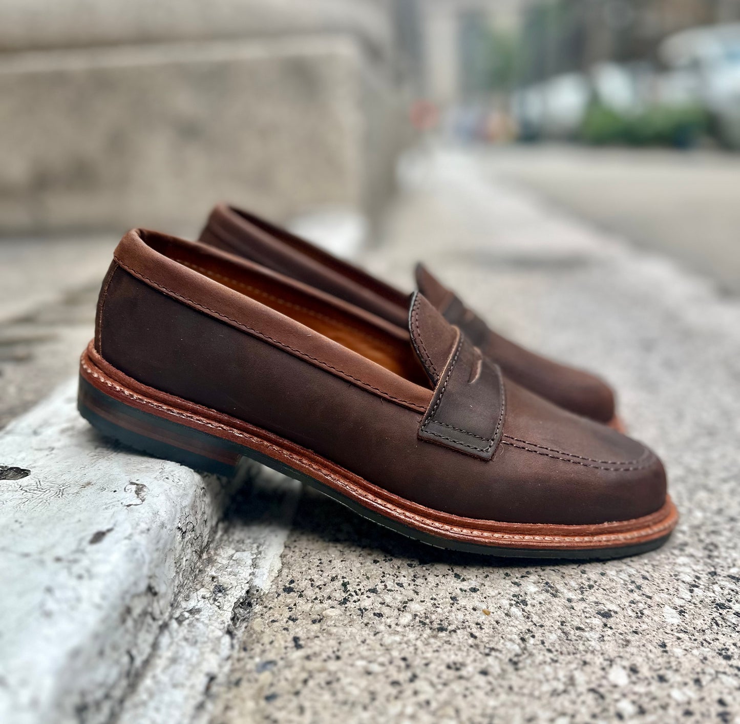The Alden Madison D2216L - Smooth Tobacco Chamois Penny Loafers, featuring an antique edge and visible sole, sit on a concrete surface with a slightly blurred urban backdrop that highlights their stitch detailing and tobacco smooth finish.
