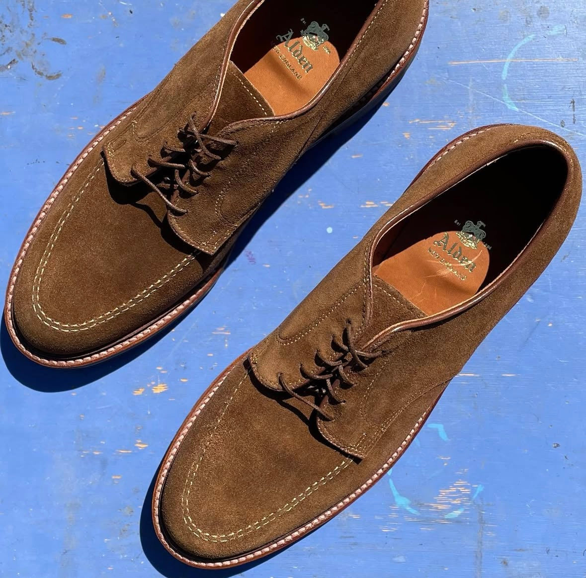 Alden Madisons 702 Mocc Toe Blucher in snuff suede, featuring white stitching and leather soles, is showcased on a textured blue surface in bright sunlight.