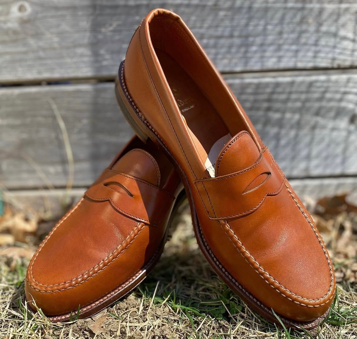 A pair of Alden Madison 983 - Leisure Hand Sewn Burnish Tan Calfskin loafers with leather outsole is displayed outdoors on grass and dry leaves, one shoe upright and the other leaning, with a wooden fence in the background.