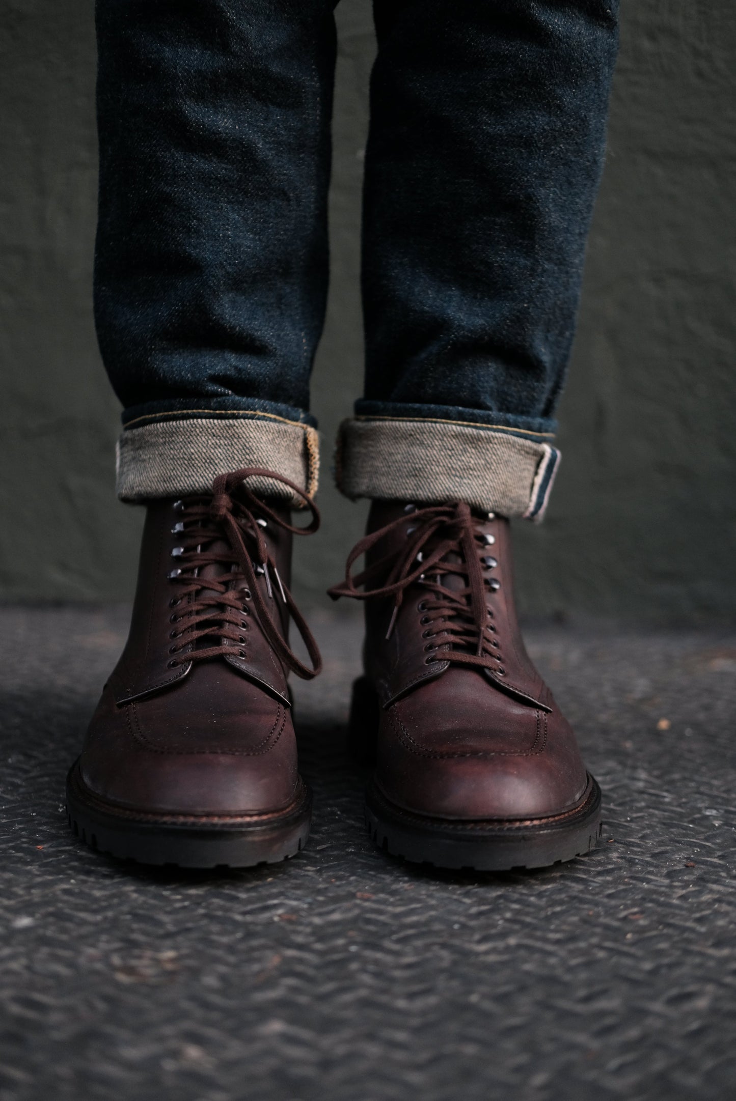 A person in cuffed dark blue jeans and Alden Madisons 404 - Indy Boot in Kudu (Oiled CXL) with a Vibram Lug Outsole stands on a textured surface, set against a plain dark wall.