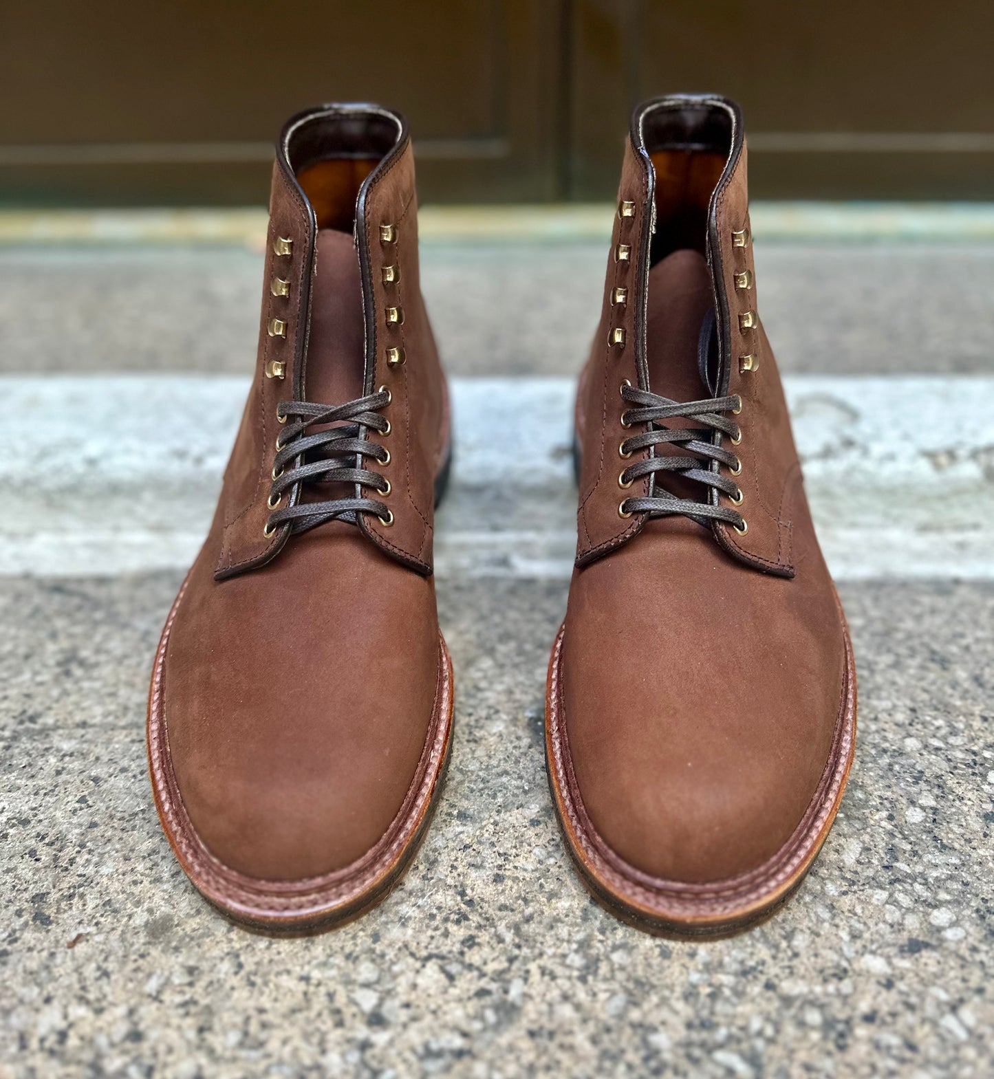 The Alden Madison 86032H Smooth Tobacco Chamois Plain Toe Boots, crafted with brown leather and metal eyelets, feature crepe soles and a polished finish. These robust boots are displayed side by side on a textured stone surface.
