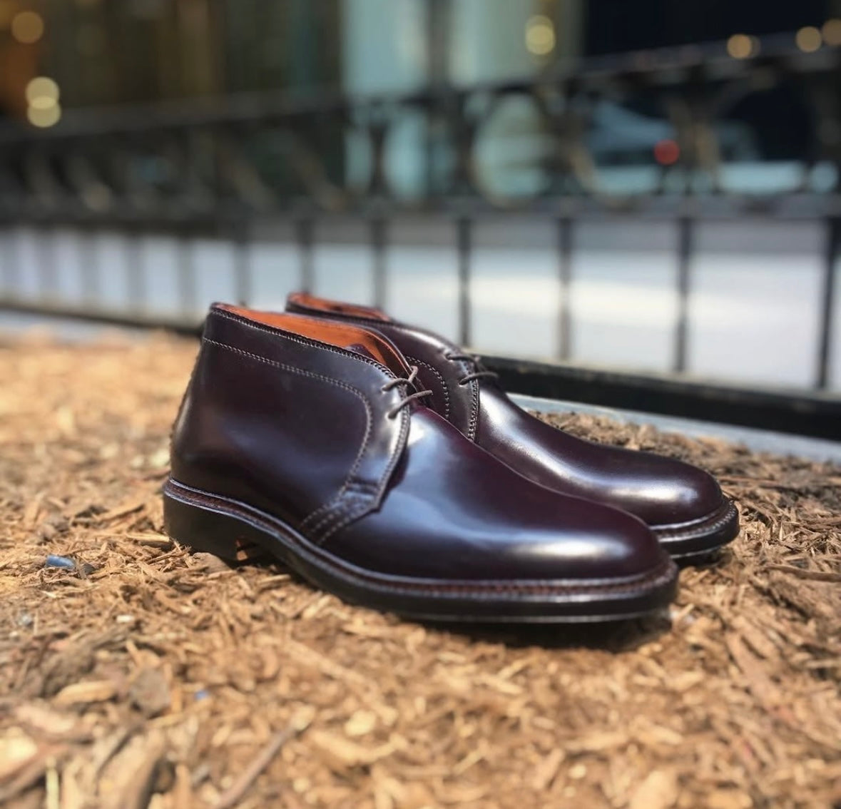 The Alden Madison 1339 Chukka Boot in Color 8 Shell Cordovan, crafted on the Barrie Last with laces, is displayed outdoors on wood mulch, with a blurred black fence and buildings in the background.