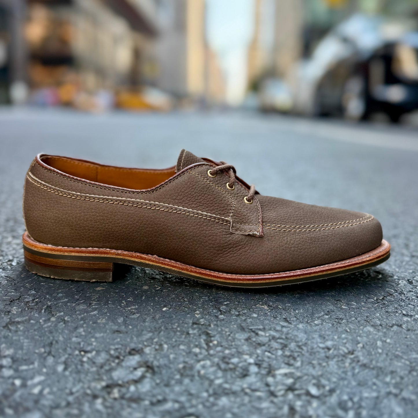 The Alden Madison 7351 - Ivy Blucher in clay nubuck, featuring visible stitching and a blucher pattern, is displayed on an asphalt street with a blurred city backdrop.