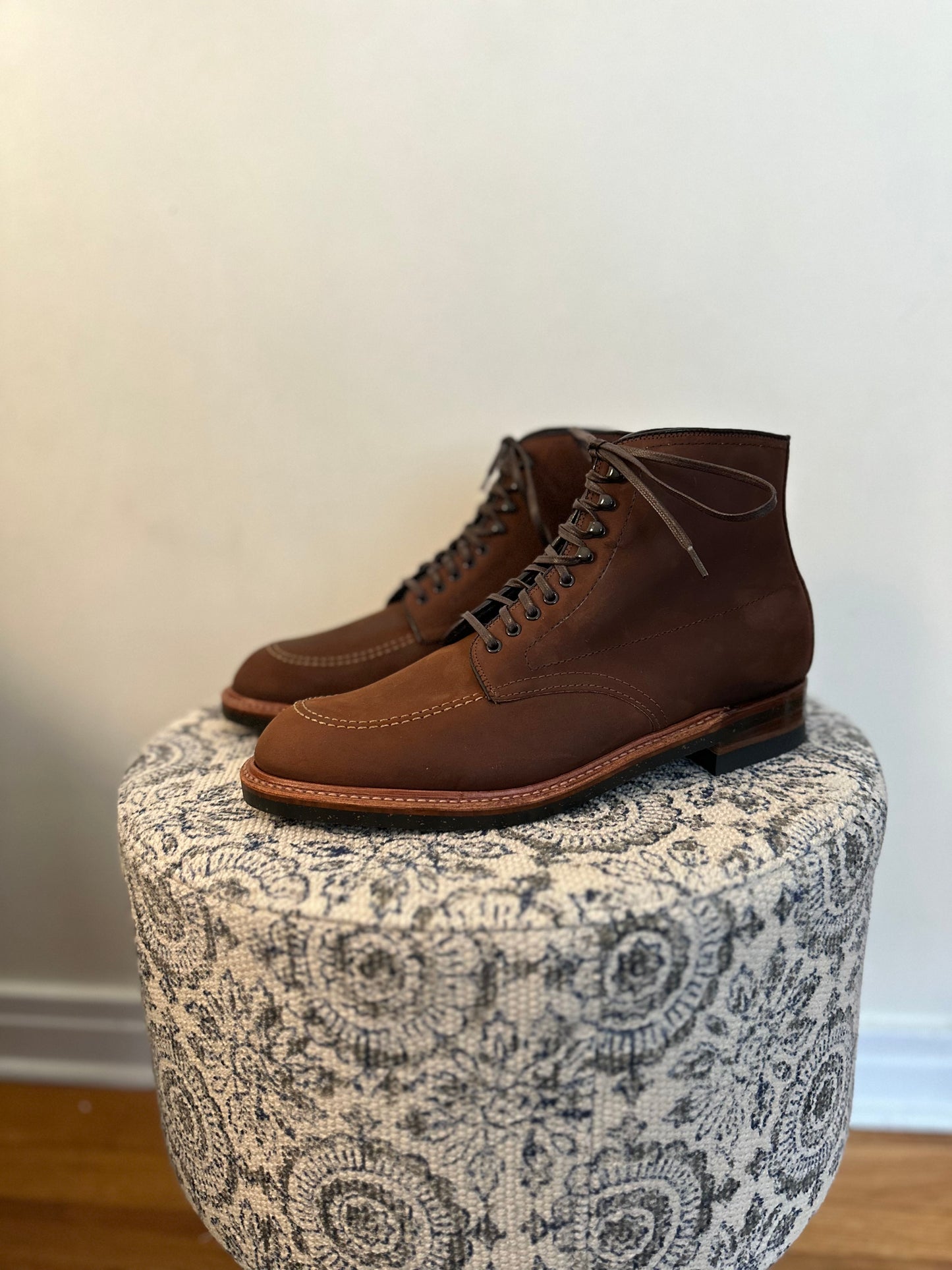 A pair of Alden Madisons D2909H - Indy Boots in Tobacco Smooth Chamois sits on a patterned, round ottoman. The brown leather and laces contrast strikingly against the plain, light-colored wall behind them.