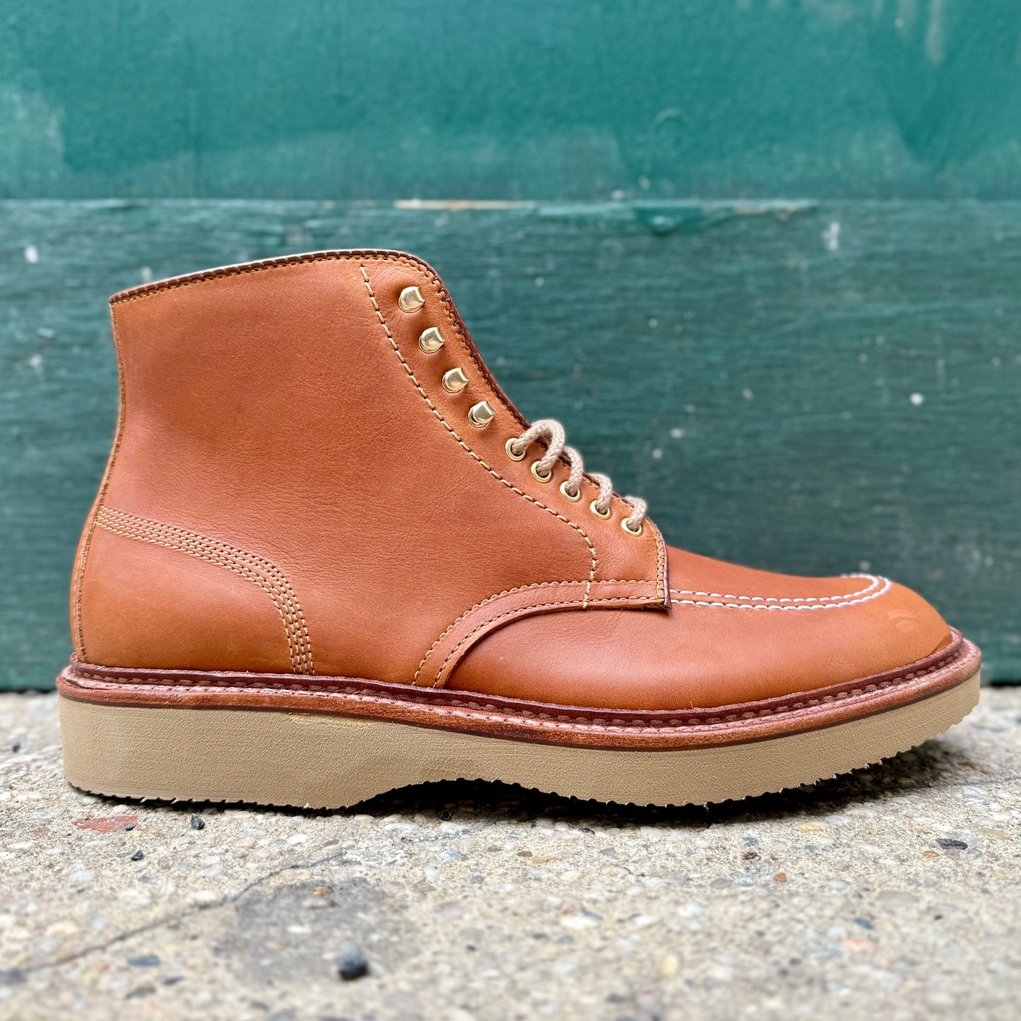 The Sherwood Indy Boot (40311H) from Alden Madison is a single tan leather boot made of saddle calf leather, featuring white stitching, tan laces with brass eyelets, and a light brown rubber sole. Its set on rough concrete with a green background.
