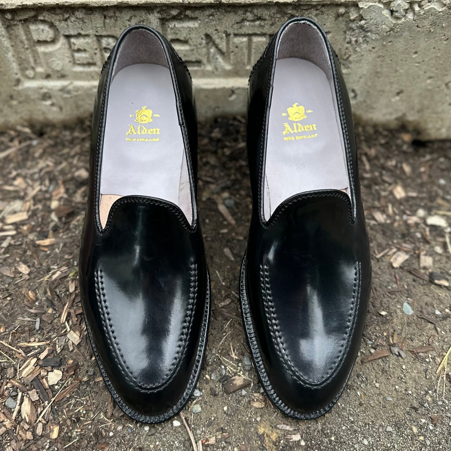A pair of D2117 - Vanderbilt Slip On loafers in Black Shell Cordovan by Alden Madison, with stitching detail and yellow brand text inside. The shoes are placed on a textured surface near a visible concrete border.