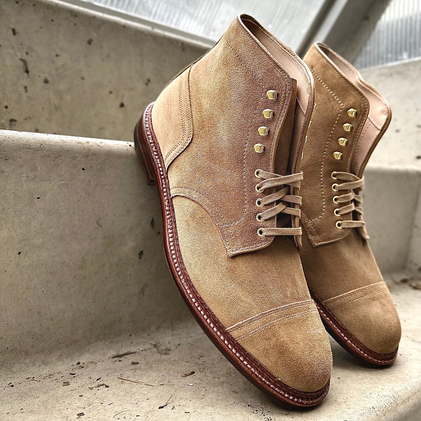 Alden Madisons D2685H - 2/2 Cap Toe Boot in Tan Suede, with brown soles and beige laces, rests on concrete steps. Crafted on the Barrie last with brass eyelets and a textured finish, its perfectly positioned under soft lighting.