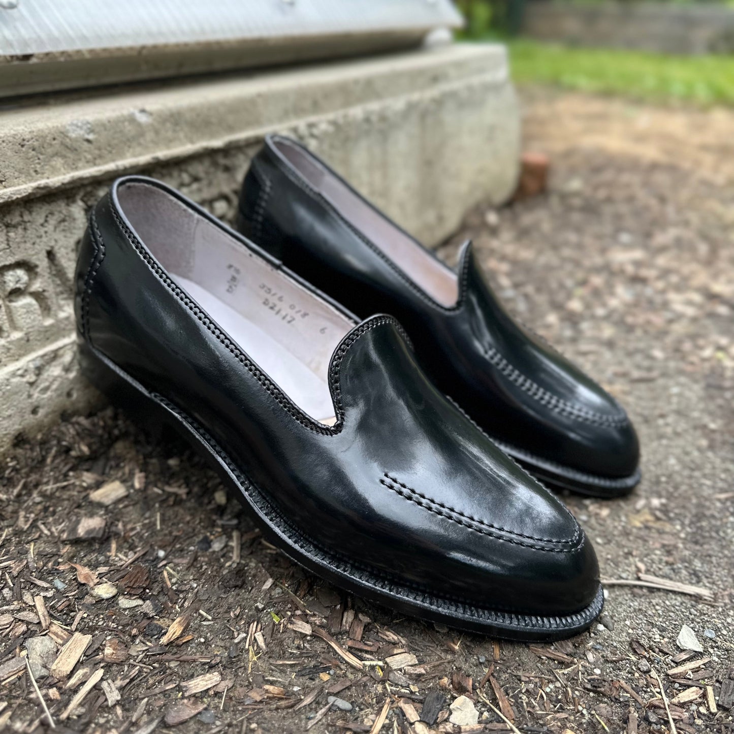 Set against a textured ground near a concrete step, the Alden Madison D2117 - Vanderbilt Slip On in Black Shell Cordovan shoes feature polished leather with a stitched design and Single Oak Leather Sole, amidst partially visible greenery, exuding timeless elegance.