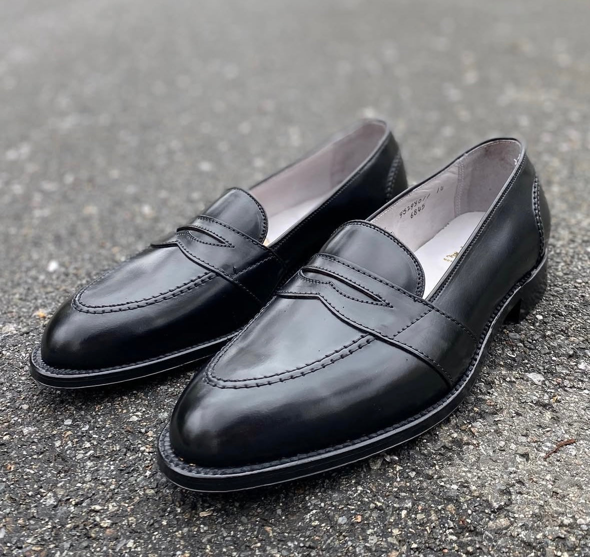 Alden Madisons 6845 Full Strap Loafer in Black Shell Cordovan, featuring stitching detail, is shown side by side on rough gray asphalt.