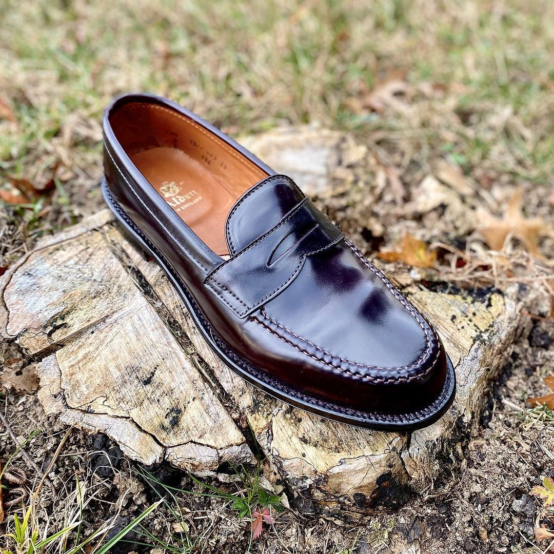 A single Alden Madison 986 - Leisure Hand Sewn loafer in Color 8 Shell Cordovan, with fine stitching and a subtle shine, featuring a luxurious leather sole, rests on a weathered tree stump amid grass and leaves.